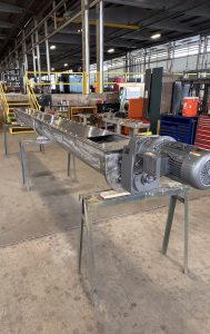 A long stainless steel conveyor system with a motor is supported by sawhorses inside an industrial workshop, with tools and equipment visible in the background.