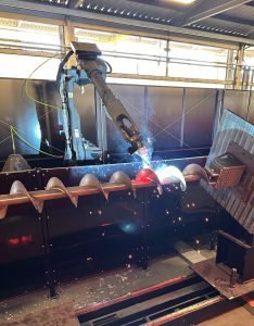 A robotic arm welds a large metal screw conveyor with optimized screw conveyor pitch in an industrial facility, sparks flying at the welding point.
