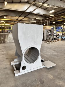 Large, gray, industrial metal hopper with a circular opening sits on a warehouse floor surrounded by shelves and sandblasting equipment, showcasing ISC’s expert Shop Services.