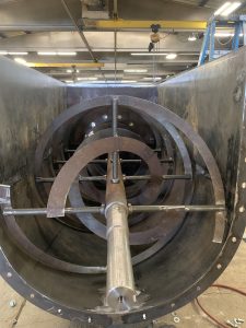 Large cylindrical metal structure with concentric rings and a central shaft, showcasing precise screw conveyor pitch and flight design, inside an industrial workshop. Tools and machinery are visible in the background.