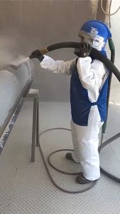 Wearing protective gear and a helmet, a person uses a large hose for sandblasting to clean a metal surface in an ISC industrial setting.