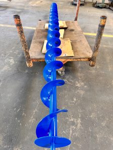 A blue metal auger with tailored flight design is positioned horizontally on a worn metal frame in an industrial workshop setting, optimizing conveyor performance.