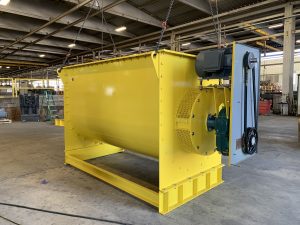 A large yellow industrial ribbon mixer with a motor and belt drive, suspended by chains inside a spacious factory, ensures efficient mixing for consistent results in demanding production environments.