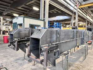 Large metal industrial components, including ISC Bucket Elevators for vertical conveying, are assembled on stands inside a spacious factory with high ceilings and various equipment in the background.