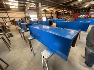 Blue metal components, including ISC Bucket Elevators, are positioned on stands inside an industrial workshop with machinery and other equipment visible in the background, showcasing reliable solutions for vertical conveying needs.