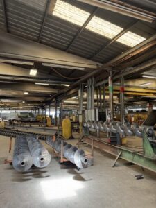 Large metal augers are lined up inside a spacious industrial warehouse with a high ceiling, exposed beams, and various machinery in the background—an impressive testament to ISC’s expertise in screw conveyor manufacturing and custom screw conveyors.