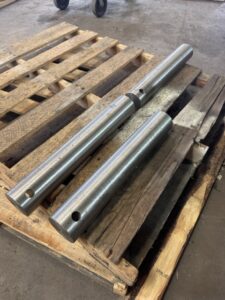 Two cylindrical metal rods with holes at each end are placed parallel on a wooden pallet in an industrial setting, suggesting components ready for assembly in a custom screw conveyors system. Their sturdy design and precision manufacture reflect principles found in advanced screw conveyor design, making them ideal for use in demanding ISC processes.
