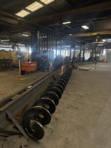Large metal auger screw lies on the floor in an industrial workshop, showcasing the precision of custom screw conveyors from ISC, with machinery and equipment visible in the background.