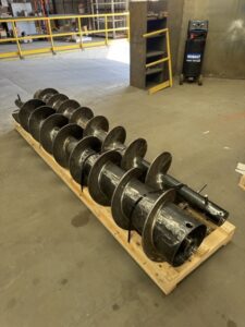 Large metal auger drill bit rests on a wooden pallet in an industrial workshop, showcasing the precision of screw conveyor fabrication; equipment and railings are visible in the background, reflecting ISC’s expertise in custom screw conveyors.