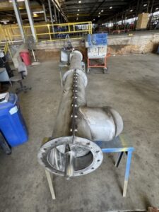 Large industrial metal pipe assembly with bolts on a stand in a warehouse, surrounded by tools, bins, and equipment in the background—ideal for ISC applications requiring custom screw conveyors or innovative screw conveyor design.