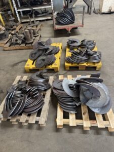 Four wooden pallets on a workshop floor hold neatly stacked curved metal parts for custom screw conveyors, with shelves and additional metal pieces—integral to advanced screw conveyor design—in the background at ISC.