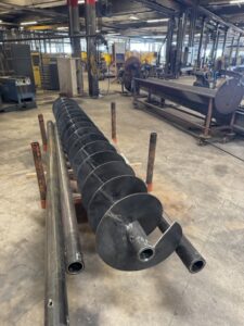 A large metal auger lies on the floor of an industrial workshop, surrounded by pipes and machinery—an example of the Not Off the Shelf quality ISC brings to custom screw conveyors crafted for specialized applications.