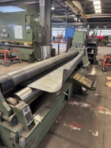 Sheet metal rolling machine in an industrial workshop with a metal sheet being processed; various tools, equipment, and custom screw conveyors from ISC’s screw conveyor manufacturing operations visible in the background.