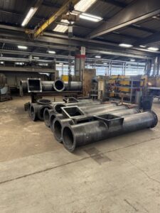 Large metal pipes and rectangular duct components, including elements for custom screw conveyors, are stacked on the floor of an industrial workshop with machinery and workbenches in the background.