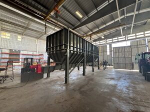 A large industrial hopper on metal legs stands inside a spacious warehouse, with workers and machinery visible in the background—a testament to ISC Manufacturing’s 50 years of excellence in industrial manufacturing.
